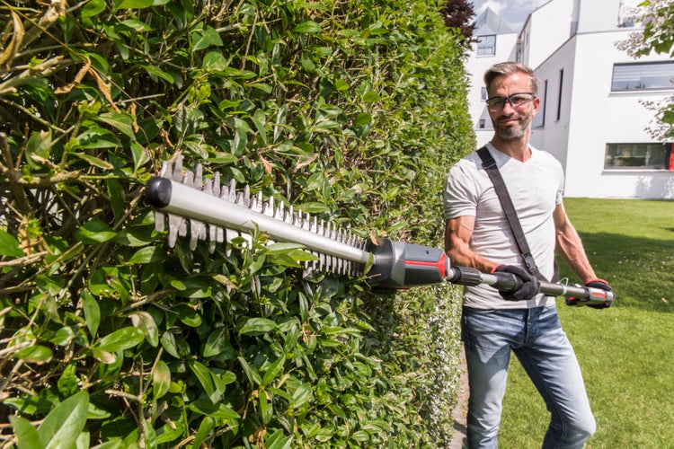 Ein Mann schneidet eine hohe grüne Hecke mit einer Teleskop-Heckenschere in einem hellen Garten.