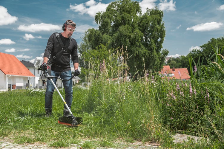 Ein Mann mäht mit einer motorbetriebenen Motorsense hohes Gras im Garten und trägt dabei Schutzkleidung.
