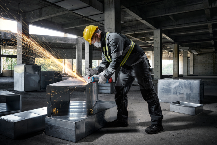 Handwerker bearbeitet Lüftungskanal mit Winkelschleifer auf Baustelle, trägt Schutzkleidung, Helm und Maske bei Funkenflug.