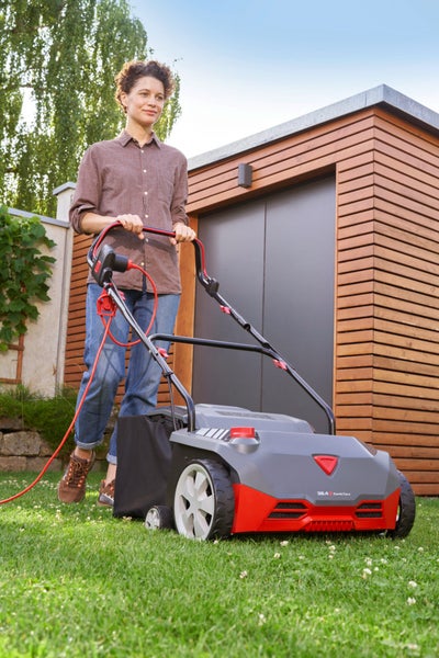 Eine Frau bedient einen Elektro-Vertikutierer auf einer Rasenfläche vor einem modernen Gartenhaus.