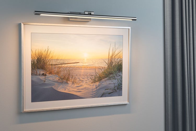 Bilderleuchte aus Metall in Chrom-Optik montiert über einem gerahmten Strandbild an einer grauen Wand.