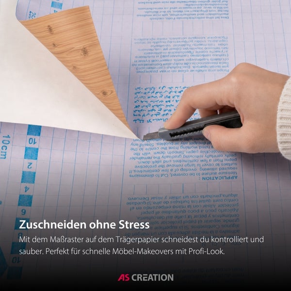 Eine Hand schneidet eine Klebefolie in Holzoptik mit einem Cuttermesser entlang des Rasters auf dem Trägerpapier zu. AS Creation Logo.