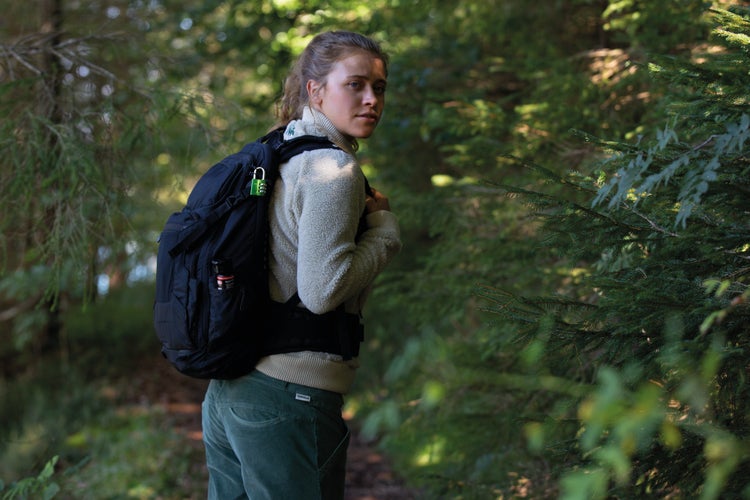 Frau wandert im Wald mit schwarzem Rucksack, gesichert durch ein grünes Zahlenschloss und ausgestattet mit einem Sicherheitsspray.