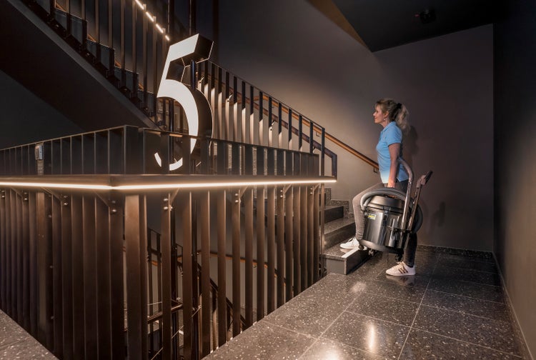 Eine Frau trägt einen Kärcher Nass- und Trockensauger eine Treppe in einem modernen Gebäude mit dunklen Wänden und beleuchteten Geländern hinauf.
