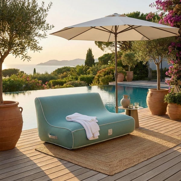 Türkisfarbenes Outdoor-Loungesofa mit weißer Paspelierung auf einer hellen Holzterrasse am Pool, mit Juteteppich, Sonnenschirm und Terrakottatöpfen.