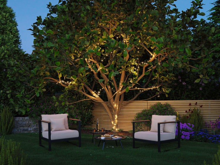 Beleuchteter Gartenbereich mit zwei Sesseln und einem Tisch unter einem Baum mit Lichterketten.