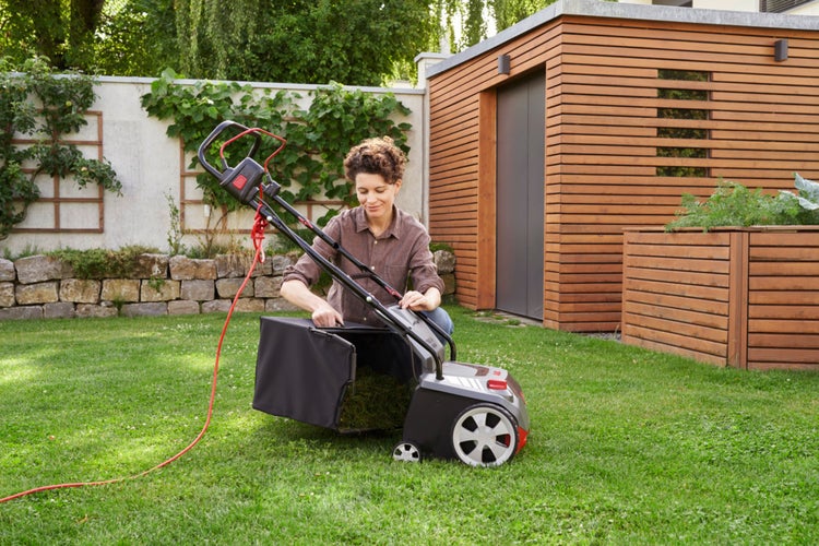 Eine Frau entleert den Fangkorb eines Elektro-Vertikutierers auf einer grünen Rasenfläche im Garten, im Hintergrund ein Holzschuppen.