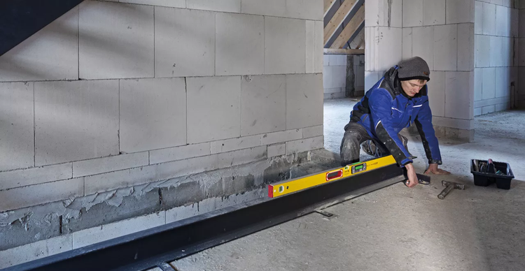 Mann prüft mit Wasserwaage und Hammer die Ausrichtung einer Bodenschiene in einem Rohbau.