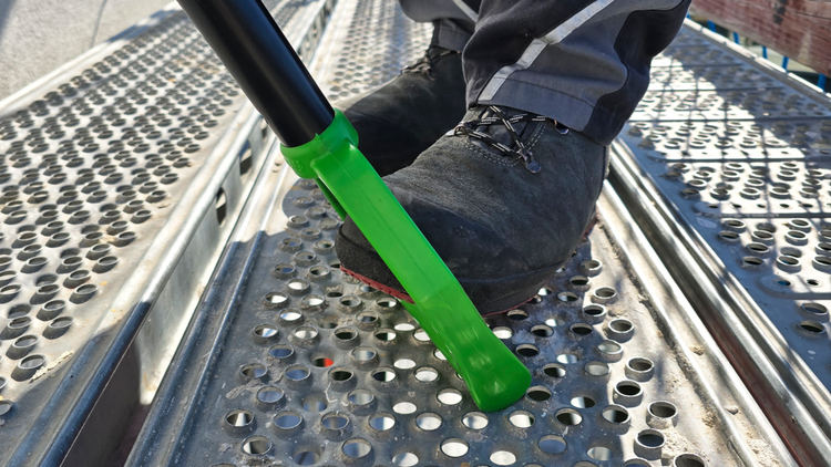 Person in Arbeitsschuhen auf einer metallenen Gerüstbohle verwendet einen Schaber mit grünem Kunststoffaufsatz.