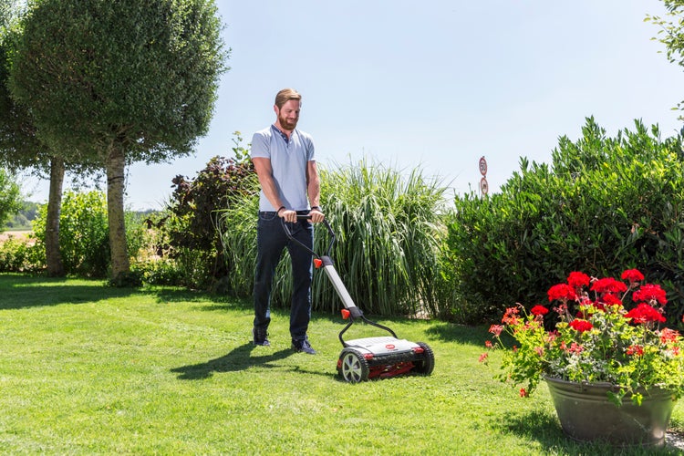 Ein Mann mäht an einem sonnigen Tag mit einem Handspindelmäher einen Gartenrasen.