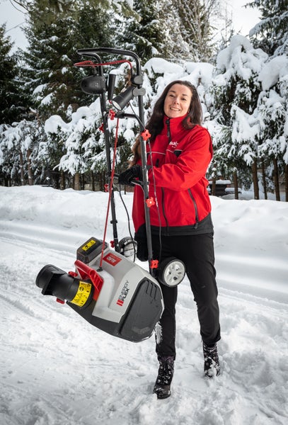 Eine Frau hält eine akkubetriebene Schneefräse AL-KO Energy Flex in einer verschneiten Winterlandschaft mit Tannenbäumen im Hintergrund.
