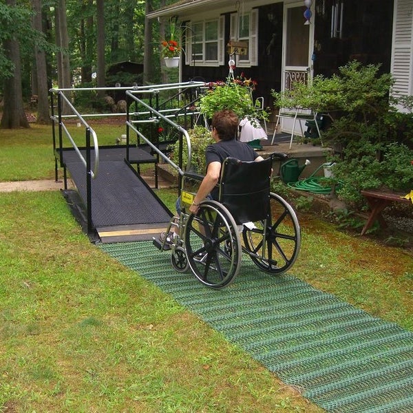 Person im Rollstuhl auf einer barrierefreien Rollstuhlrampe mit Geländer vor einem Wohnhaus und einer temporären Gehwegmatte im Garten