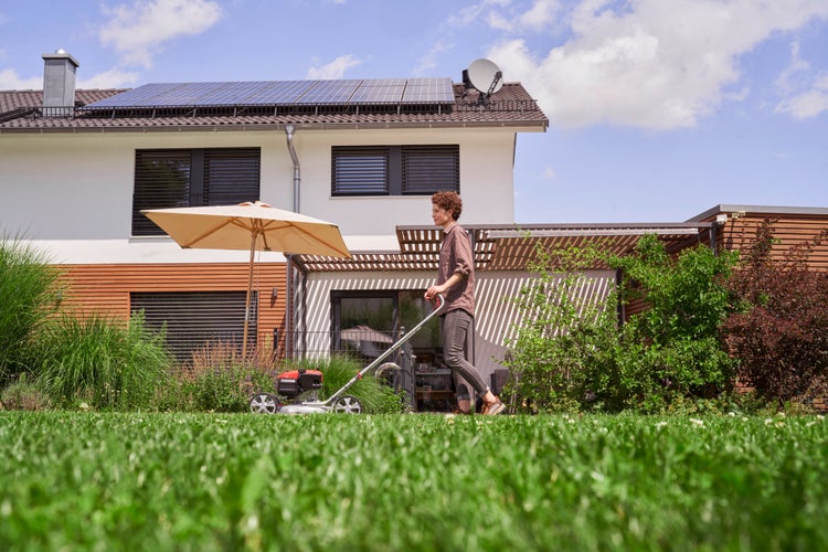 Eine Person mäht den Rasen im Garten vor einem modernen Wohnhaus mit Solaranlage und Terrasse unter einem Sonnenschirm.