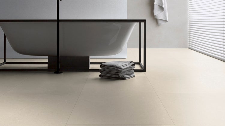 Modernes, helles Badezimmer mit großformatigen Bodenfliesen, einer freistehenden Badewanne in einem Rahmen und einem Stapel Handtücher.