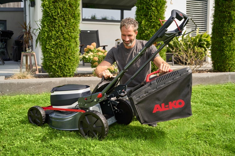 Ein Mann setzt den Grasfangkorb an einen AL-KO Rasenmäher auf einer Rasenfläche im Garten ein.