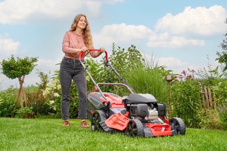 Eine Frau mäht einen Rasen mit einem Benzinrasenmäher in einem Garten mit Pflanzen und Blumen an einem sonnigen Tag.