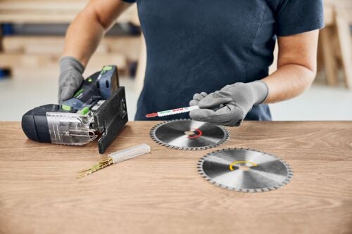 Eine Person mit Handschuhen hält ein Stichsägeblatt über Kreissägeblätter, neben einer Stichsäge auf einem Tisch