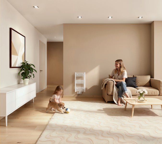 Wohnzimmer mit Frau, Kind und Hund auf einem Teppich, an der Wand ein Badheizkörper, helle Einrichtung mit Parkettboden und einem weißen Sideboard.