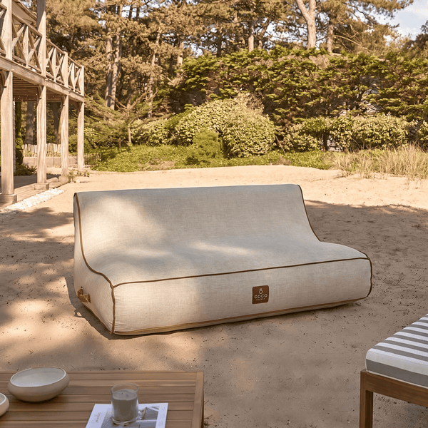 Outdoor-Lounge-Sofa aus hellem Textil mit brauner Paspelierung auf Sand stehend, im Hintergrund Bäume und eine Holzkonstruktion.