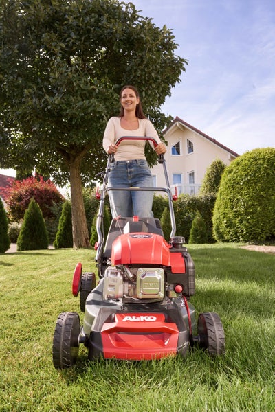 Frau beim Mähen des Rasens mit einem AL-KO Benzin-Rasenmäher in einem Garten mit Wohnhaus.