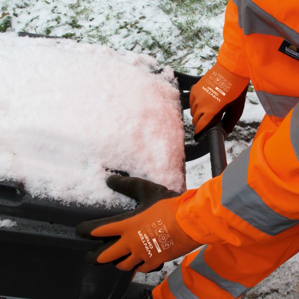 Eine Person in Arbeitskleidung mit Winter Grip Handschuhen schiebt eine schneebedeckte Schubkarre.