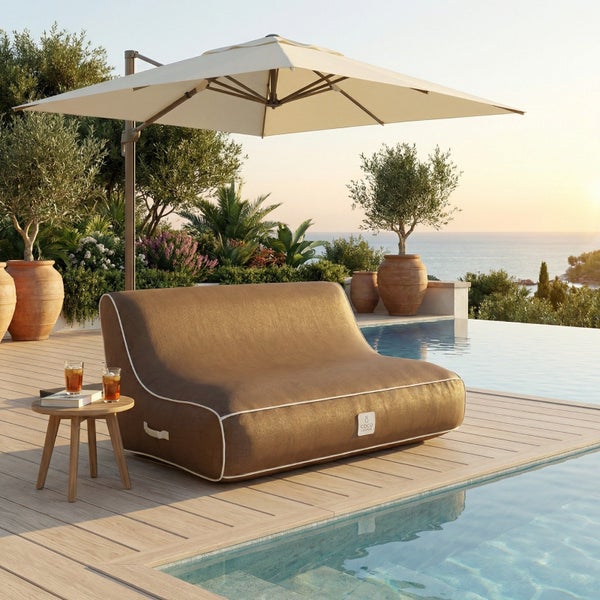 Braunes Outdoor-Sitzsack-Sofa mit weißer Biese auf einer Holzterrasse neben einem Pool unter einem großen Sonnenschirm.
