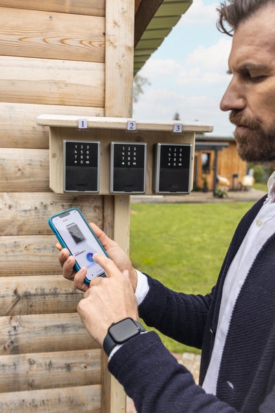 Ein Mann steuert einen von drei an einer Holzwand montierten elektronischen ABUS Schlüsselsafes über eine Smartphone App im Außenbereich.