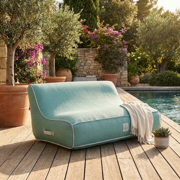 Hellgrünes Outdoor-Lounge-Sofa auf einer Holzterrasse neben einem Pool mit mediterranen Pflanzen im Hintergrund. Coco Logo.
