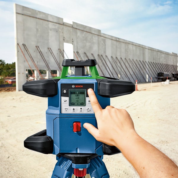 Bosch Rotationslaser auf einem Stativ auf einer Baustelle mit Betonwand im Hintergrund, ein Finger bedient das Display und die Tasten des Geräts.