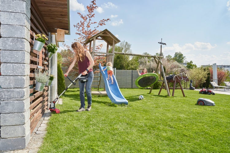 Eine Frau nutzt einen Rasentrimmer an einem Gartenhaus, während Kinder auf einem Spielplatz mit Rutsche und Schaukel im Garten spielen.