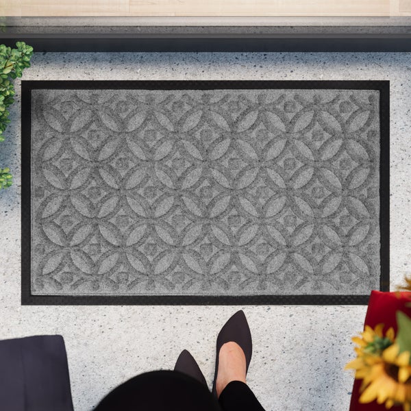 Fußmatte mit geometrischem Kreismuster und Gummirand auf einer hellen Steinterrasse vor einer Eingangstür.