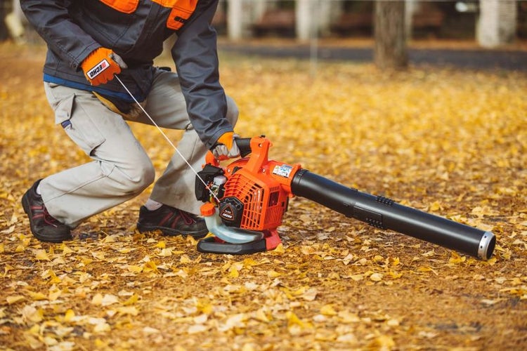 Person startet Laubbläser auf einer Fläche mit Herbstlaub.