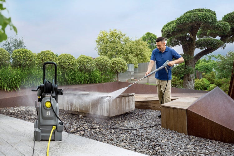 Ein Mann verwendet einen Hochdruckreiniger zur Reinigung einer Betoneinfassung in einem modernen Garten mit Kies, Pflanzen und Holzterrasse.