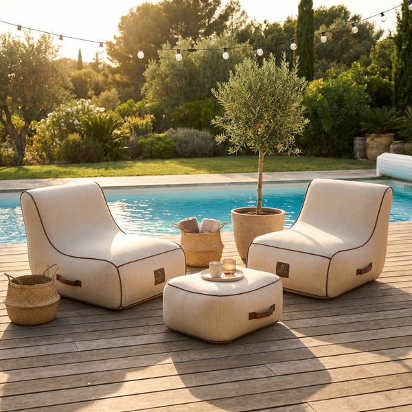 Zwei beige Gartenloungesessel und ein passender Hocker auf einem Holzdeck neben einem Pool in einem hellen, weitläufigen Garten mit Olivenbaum.
