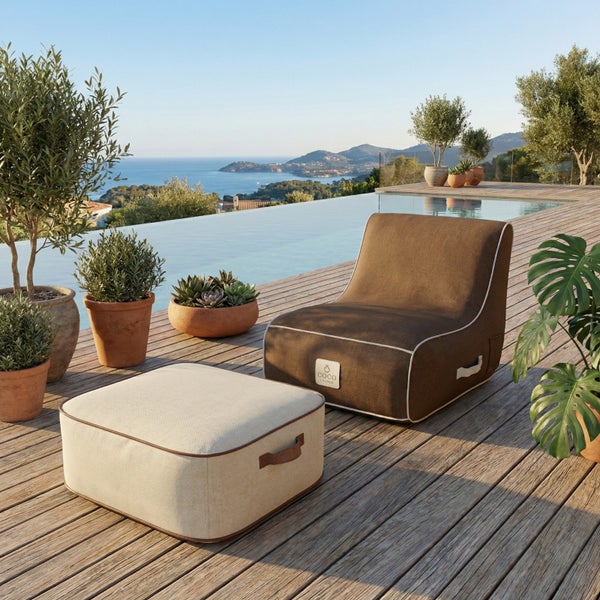 Moderner Loungesessel und Hocker aus Textilgewebe auf einer hellen Holzterrasse neben einem Pool mit Blick auf das Meer und die Küste.