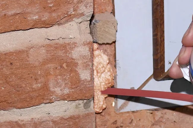 Auf dem Bild wird Bauschaum zwischen einem Ziegelstein und einem Fensterrahmen aufgetragen.
