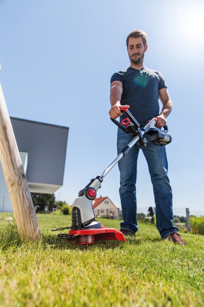 Ein Mann nutzt einen akkubetriebenen Rasentrimmer, um Kanten an einem Holzpfahl im Garten zu schneiden.