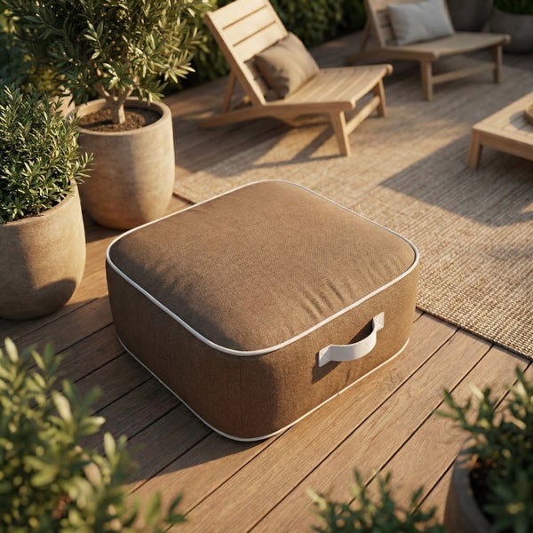 Quadratischer Outdoor-Pouf aus braunem Stoff mit weißer Paspelierung und Griff auf einer hellen Holzterrasse mit Gartenmöbeln und Pflanzen.