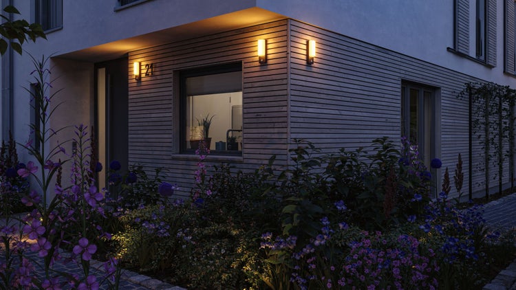 Moderne Außenbeleuchtung an einer Holzfassade über einem bepflanzten Blumenbeet mit violetten Blüten in der Abenddämmerung.