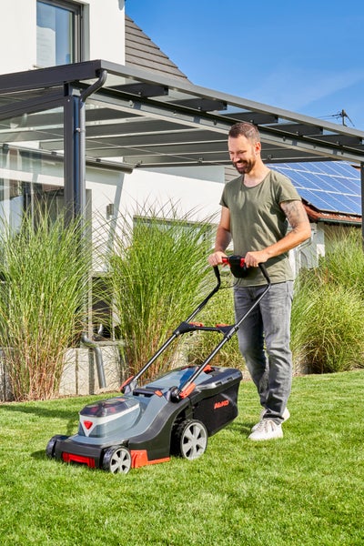 Ein Mann mäht den grünen Rasen im Garten mit einem AL-KO Akku-Rasenmäher vor einer modernen Terrasse.
