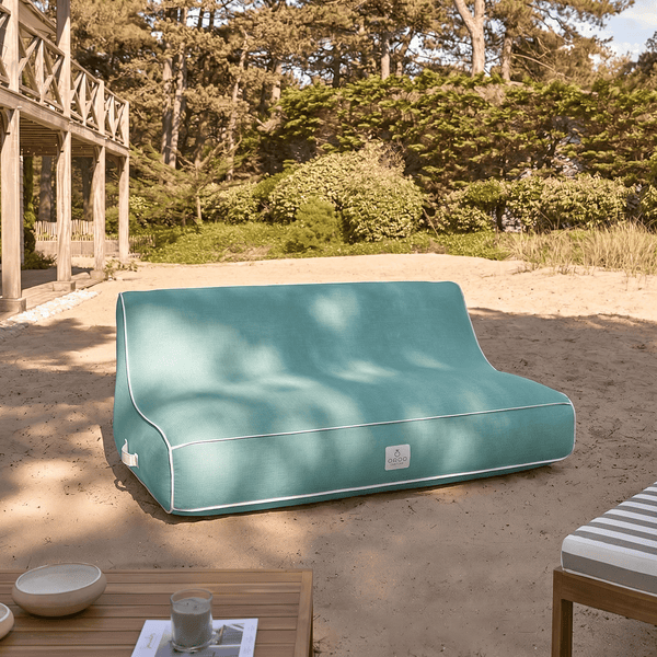 Türkisfarbenes Outdoor-Lounge-Sofa auf Sand in einem hellen Garten mit Holzterrasse und Bäumen im Hintergrund.