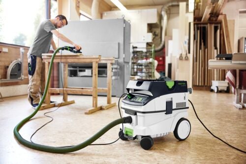 Handwerker nutzt ein Festool Absaugmobil in einer hellen Holzwerkstatt beim Bearbeiten eines Werkstücks auf einer Hobelbank.