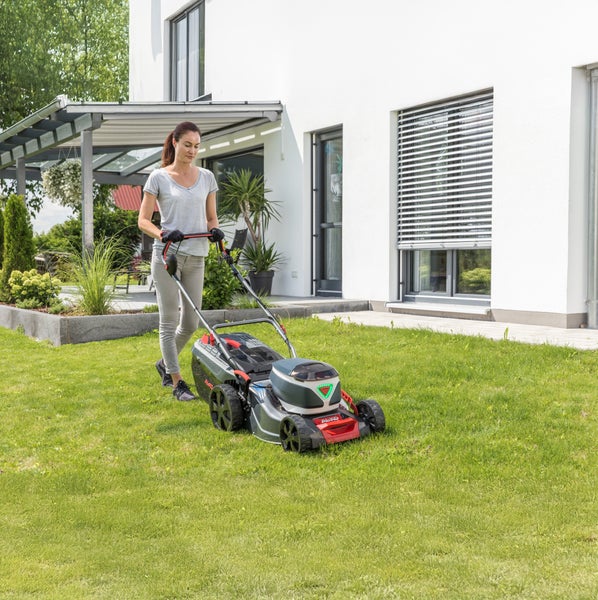 Eine Frau mäht mit einem Akku Rasenmäher der Marke AL KO den Rasen vor einem modernen Wohnhaus.