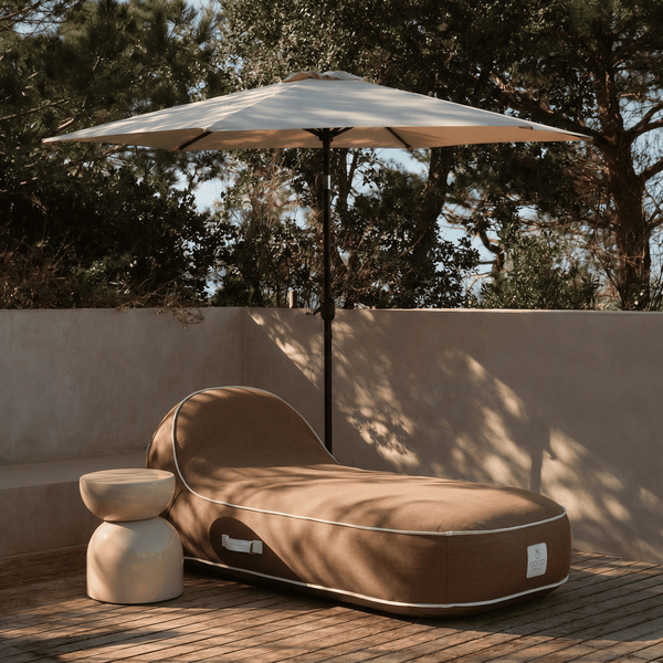 Brauner Outdoor-Sitzsack auf einer Holzterrasse neben einem Beistelltisch im Schatten eines weißen Sonnenschirms.