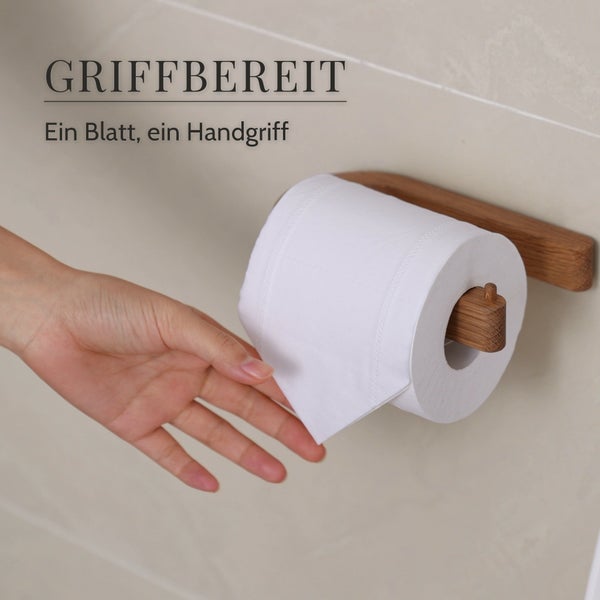 Ein hölzerner Toilettenpapierhalter an der Wand mit einer Rolle Toilettenpapier, eine Hand greift nach einem Blatt.