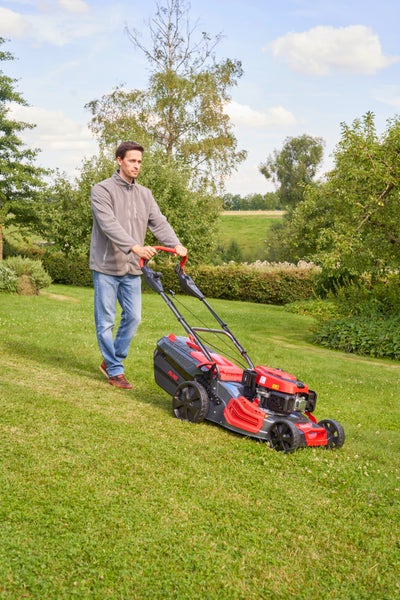 Mann beim Rasenmähen mit einem benzinbetriebenen Rasenmäher der Marke AL-KO auf einer sonnigen Gartenfläche im Freien.