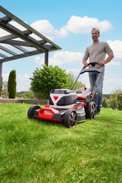 Mann mäht Rasen mit Akku-Rasenmäher im hellen, organisierten Garten mit Pergola und Sträuchern.