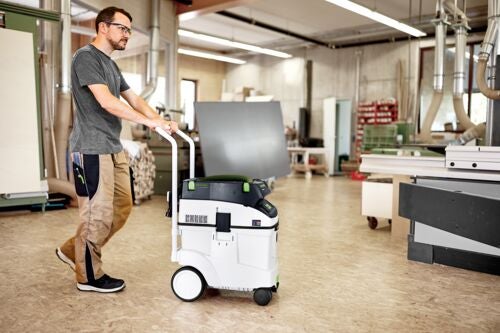 Ein Handwerker schiebt einen mobilen Werkstattsauger mit Schiebebügel durch eine helle, organisierte Werkstatt mit Holzelementen.