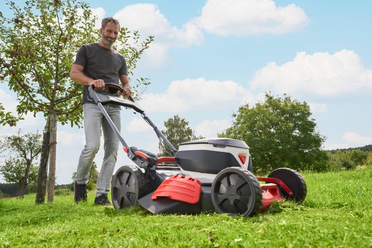 Mann mäht Gras mit einem modernen Rasenmäher in einem hellen, weitläufigen Garten mit Obstbäumen.