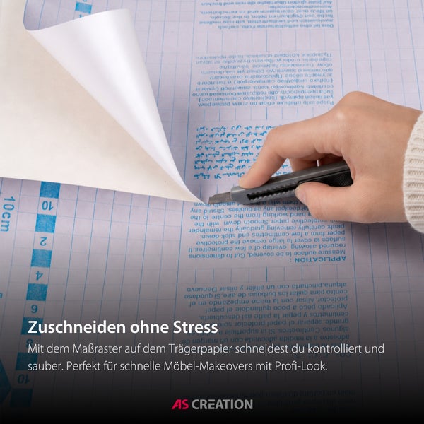 Eine Hand schneidet eine selbstklebende Folie mit einem Cuttermesser entlang des Rasters auf dem Trägerpapier zu. AS Création Logo.